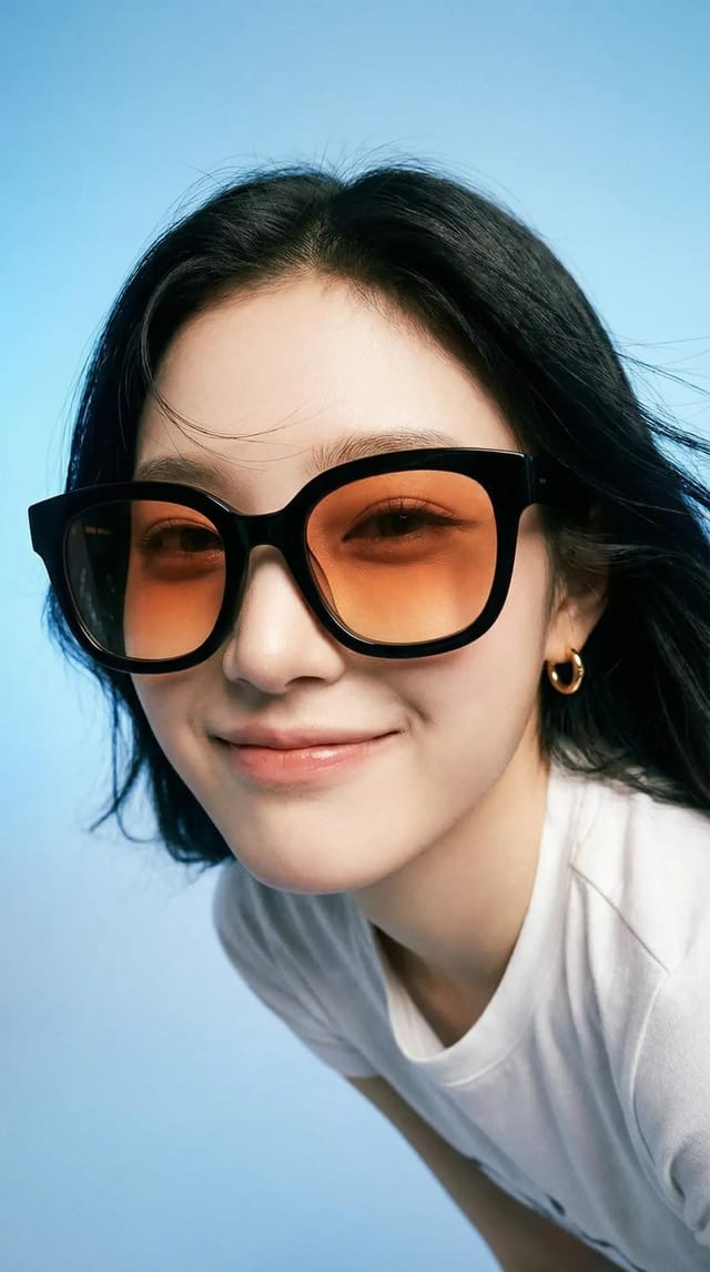 Portrait image of a smiling woman wearing oversized black sunglasses with orange lenses against a blue studio background.